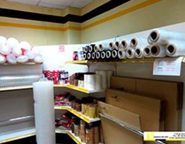 Pasillo de tienda con estantes de productos de papel, rollos de plástico y cajas de cartón en una habitación que parece un almacén.