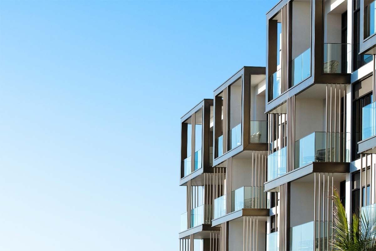 Edificio de apartamentos moderno con balcones cuadrados y barandillas de vidrio contra un cielo azul claro.