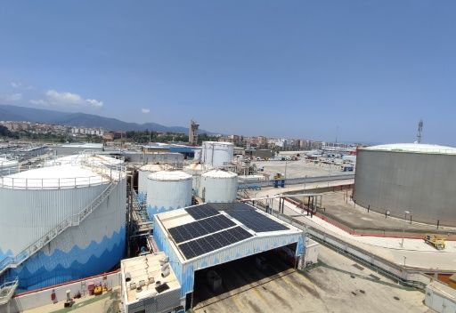 Sitio industrial con tanques de almacenamiento blancos, paneles solares en un edificio, cielo azul y una ciudad costera al fondo.