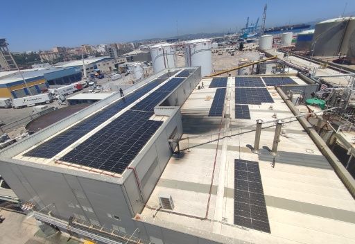 Paneles solares en el tejado de un edificio, con estructuras industriales y un cielo azul al fondo.