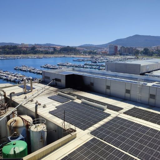 Paneles solares en el tejado de una fábrica con vistas a un puerto deportivo con barcos. Cielo azul y montañas a lo lejos.