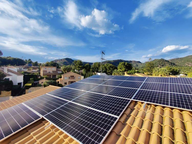 Paneles solares sobre un techo de terracota contra un cielo azul con nubes y una cadena montañosa lejana.