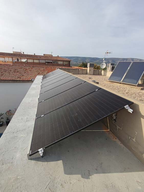 Paneles solares instalados en un tejado, orientados hacia el sol. Otros paneles solares y edificios al fondo.