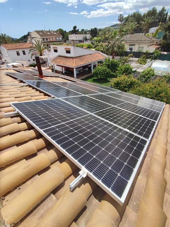 Paneles solares instalados en un techo de tejas de terracota, con casas y vegetación al fondo en un día soleado.