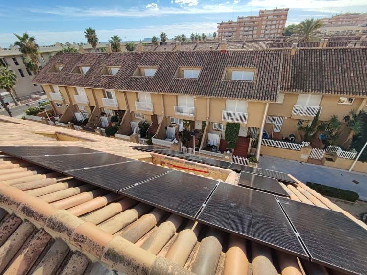 Paneles solares sobre un tejado de tejas delante de una hilera de casas adosadas de color beige con pequeños balcones.