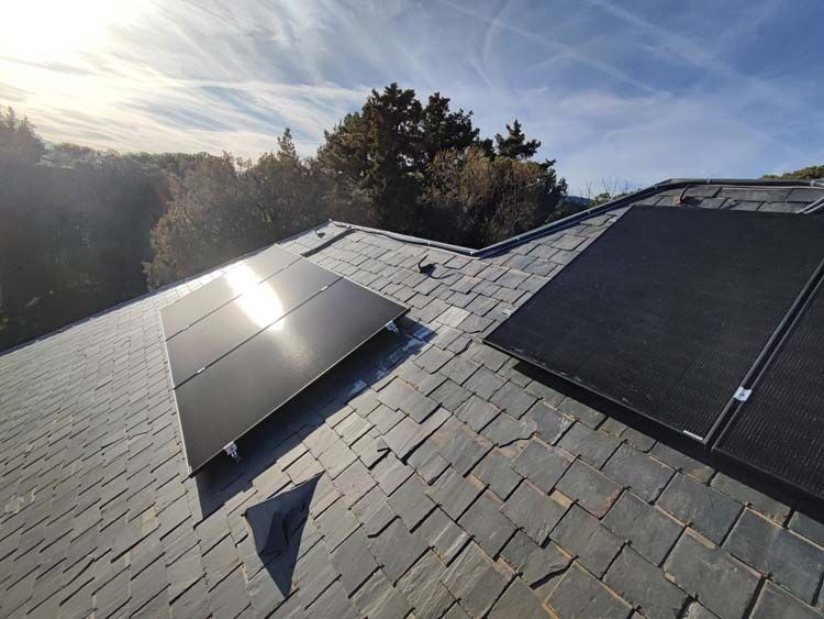 Paneles solares instalados en un techo de pizarra, contra un cielo azul con árboles visibles.
