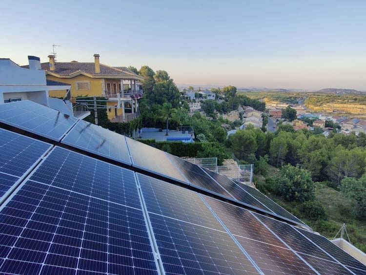 Paneles solares sobre un tejado inclinado con un pueblo al fondo. Los paneles azules reflejan el cielo y el entorno.