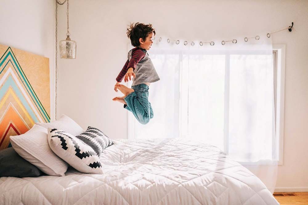 Niño saltando en la cama, dormitorio iluminado por el sol.