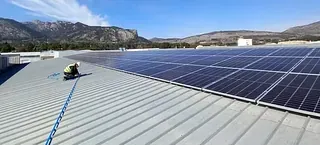 Un trabajador en un techo de metal, instalando paneles solares bajo un cielo azul claro, con montañas al fondo.