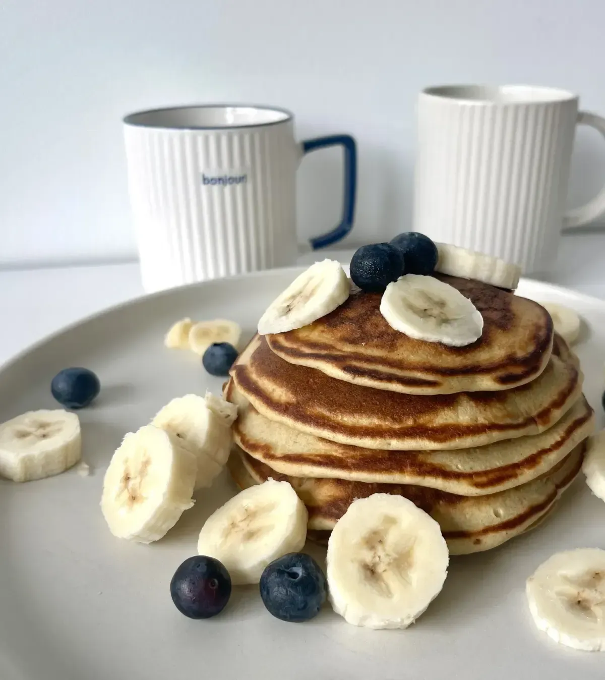 Pila de panqueques cubiertos con plátano y arándanos, con dos tazas blancas al fondo.