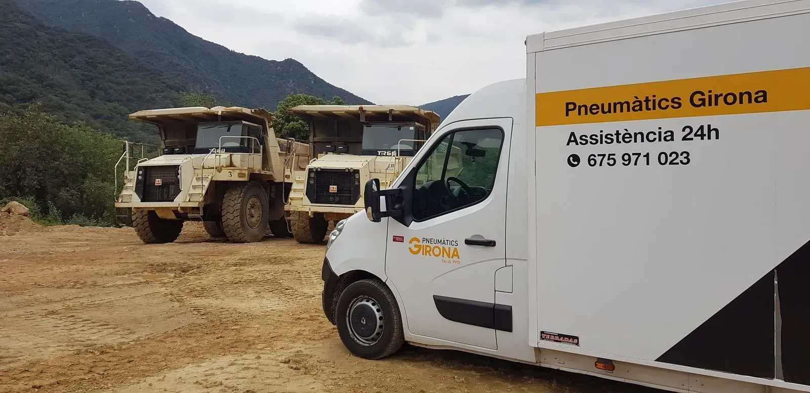 Una furgoneta blanca està aparcada al costat d'un camió de bolquet en un camp de terra.