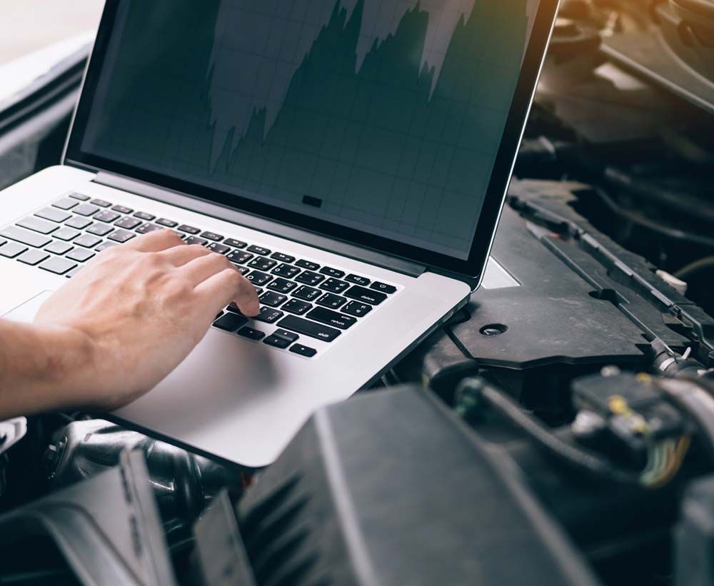 Una persona está escribiendo en una computadora portátil frente al motor de un automóvil.