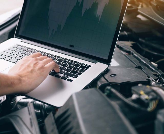 Una persona está escribiendo en una computadora portátil frente al motor de un automóvil.