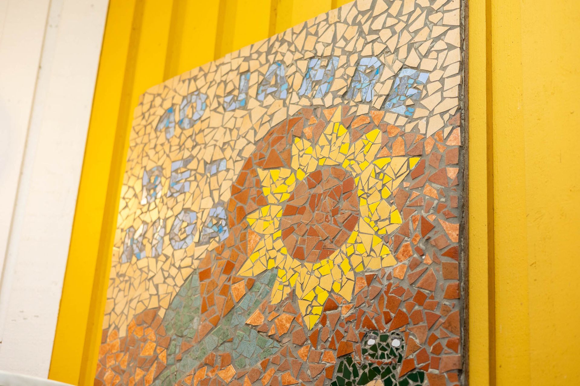 An einer gelben Wand hängt ein Mosaikgemälde einer Sonnenblume.