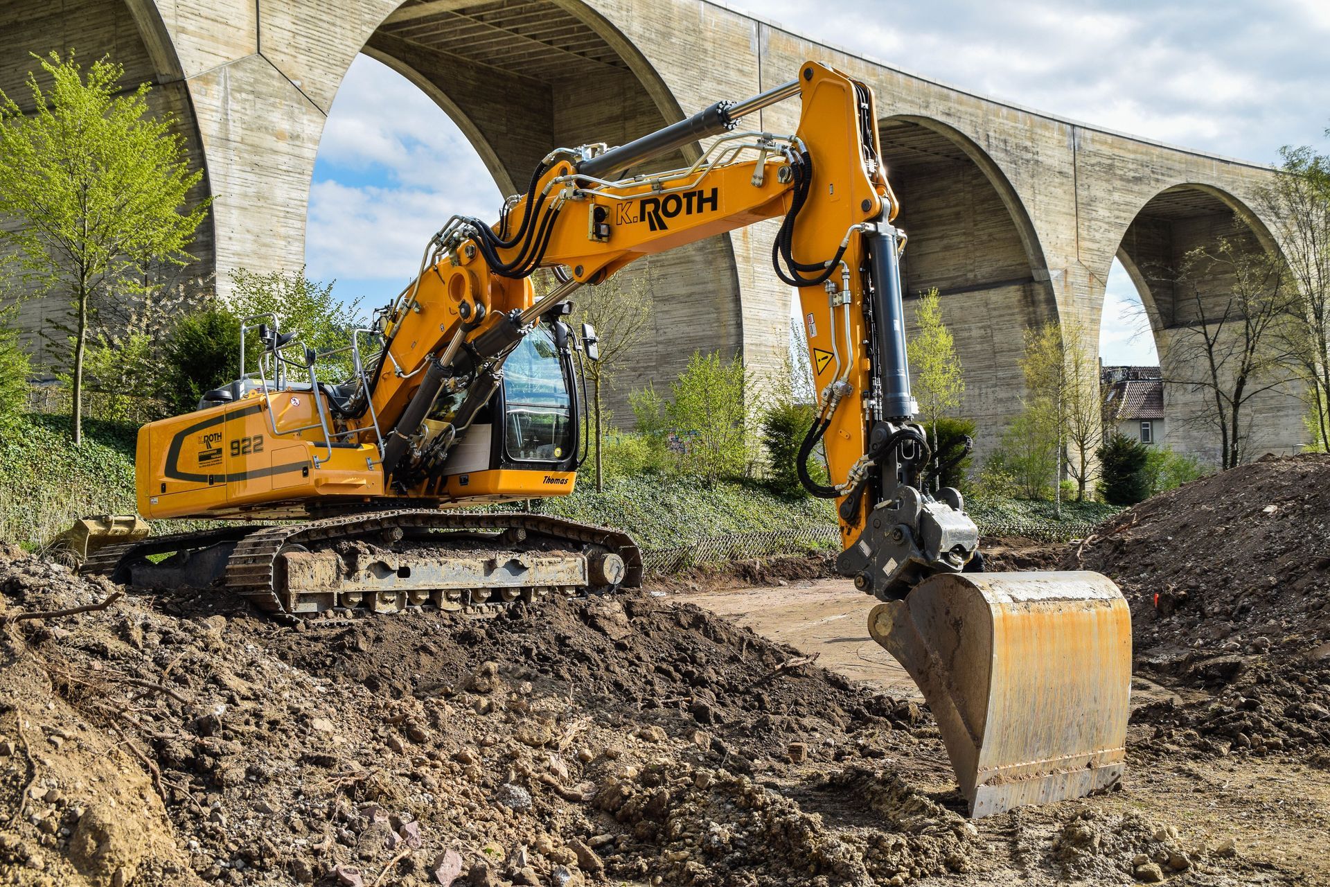 Ein gelber Bagger steht auf einem Erdhaufen vor einer Brücke.