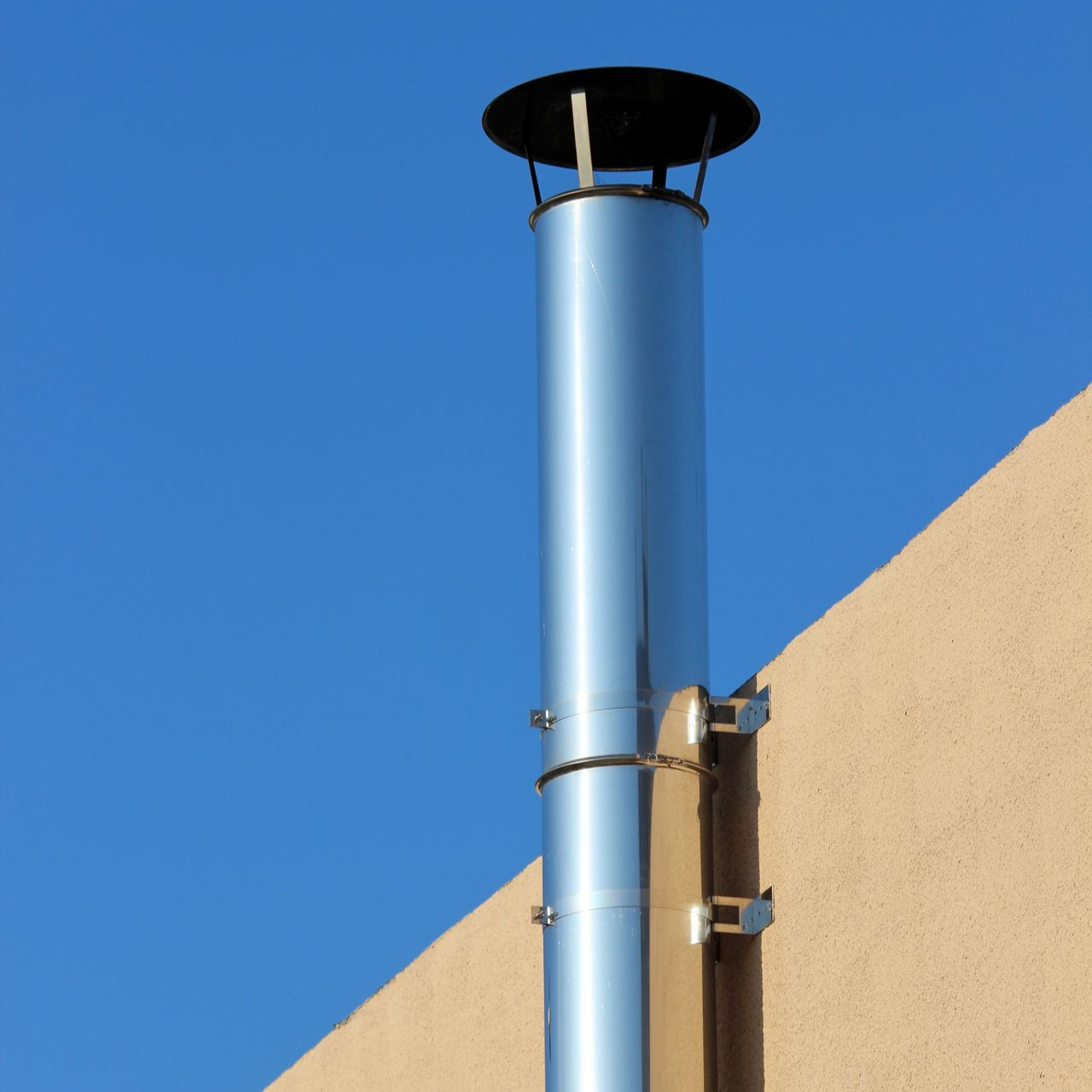 Conduit de cheminée en métal brillant fixé à un bâtiment beige sur fond de ciel bleu clair.