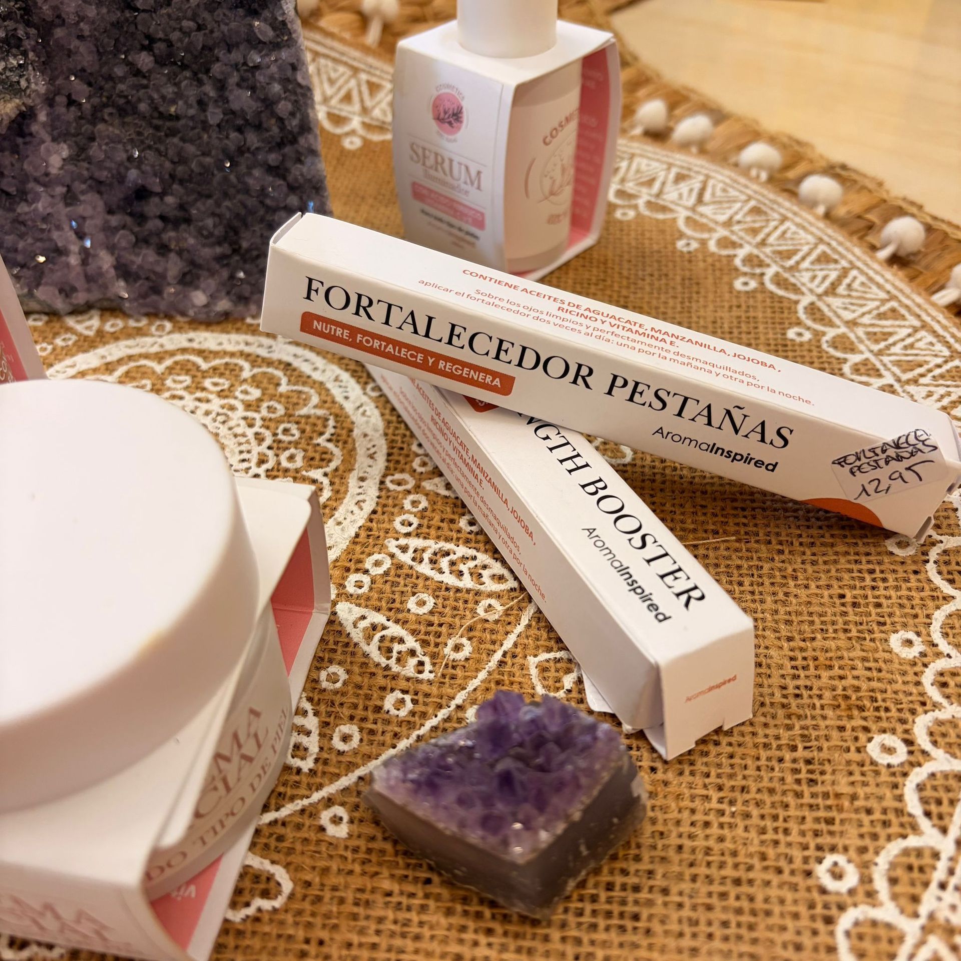 Cosméticos sobre una superficie de arpillera. Cajas de potenciador de pestañas, un frasco de sérum y cristales morados.