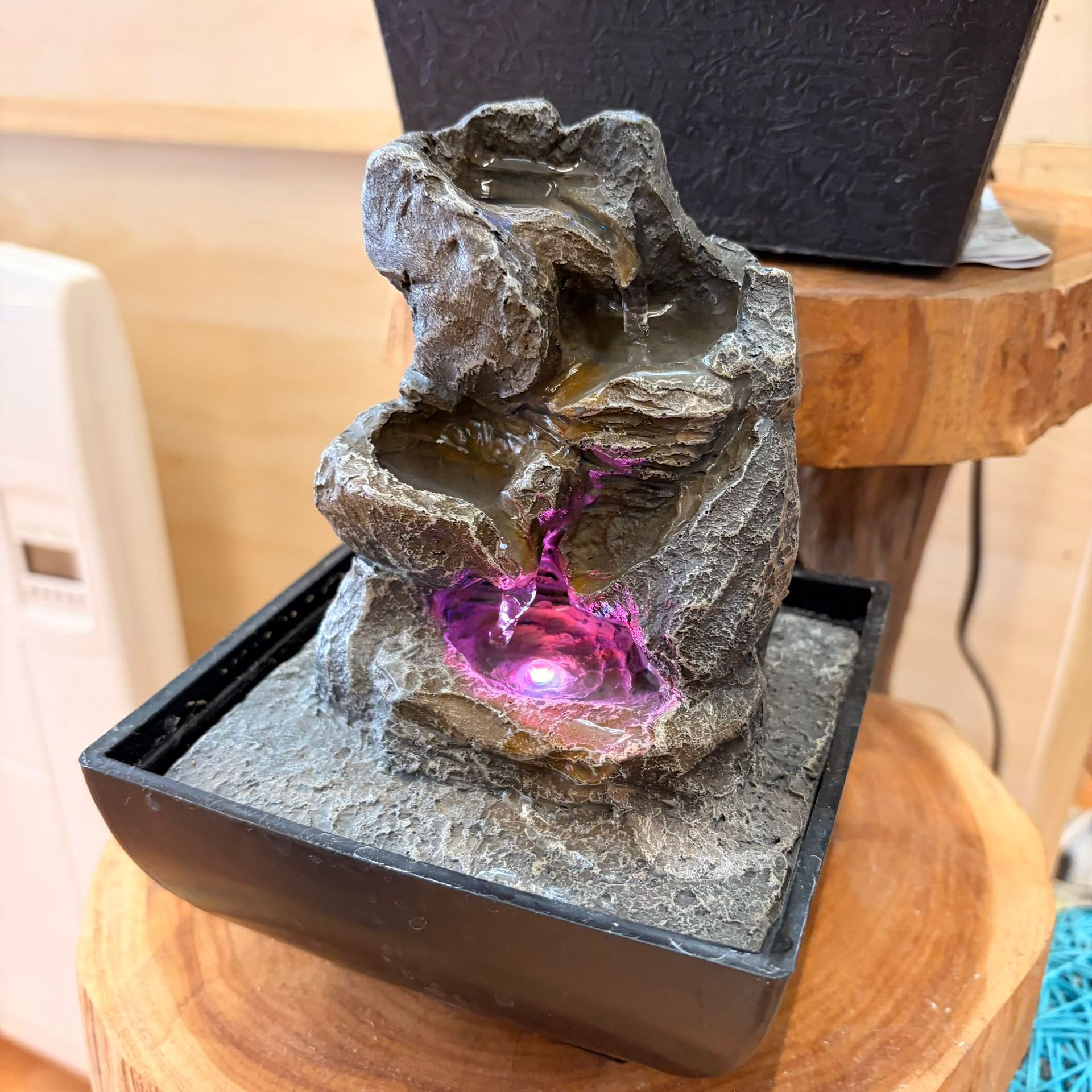 Pequeña fuente de agua de interior, diseño de roca gris con luz LED violeta, colocada sobre una superficie de madera.