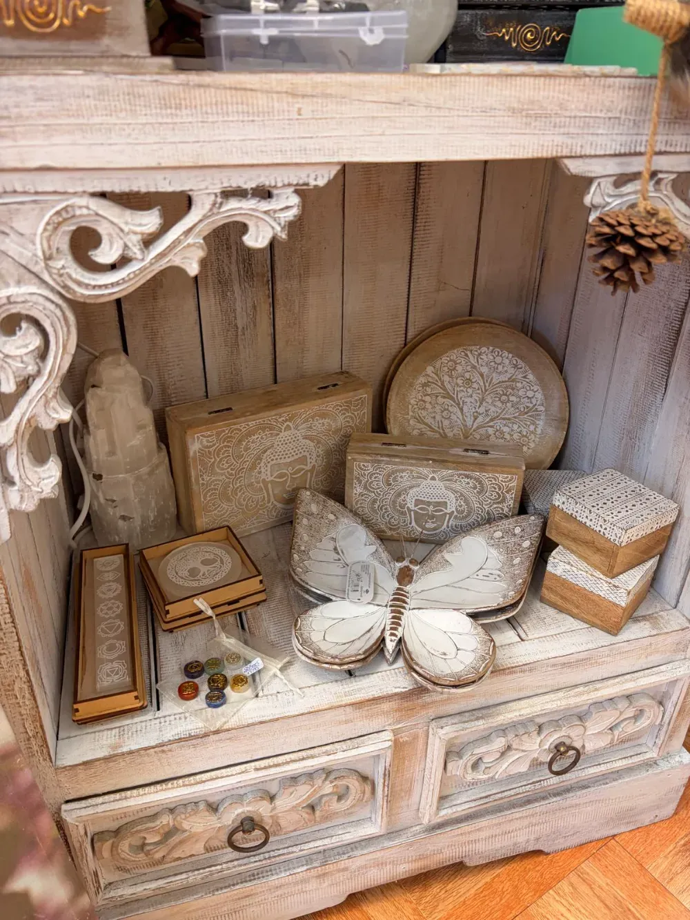 Vitrina de madera blanca envejecida que exhibe cajas decorativas, una mariposa y un plato con diseño floral.