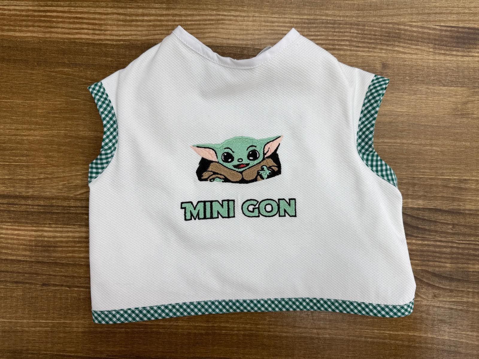 Un babero blanco con un Baby Yoda bordado está sobre una mesa de madera.