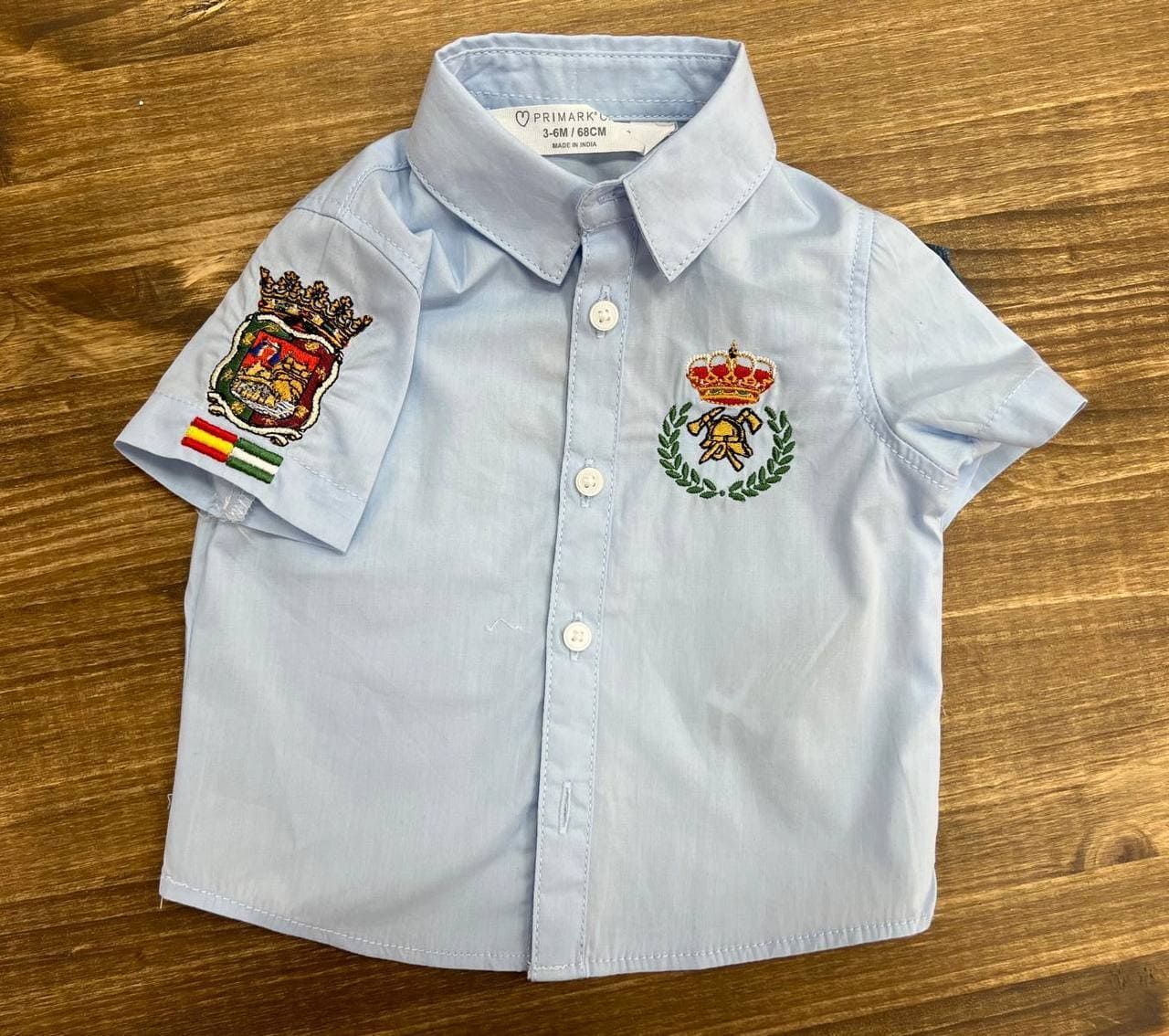 Una camisa azul claro con una corona y banderas en las mangas está sobre una mesa de madera.