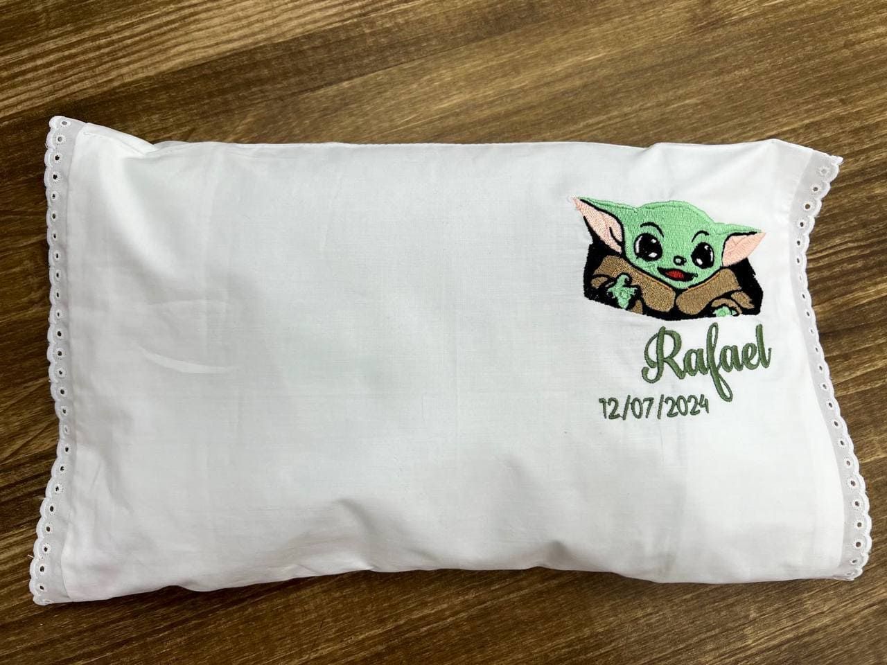 Una almohada blanca con un Baby Yoda bordado está sobre una mesa de madera.