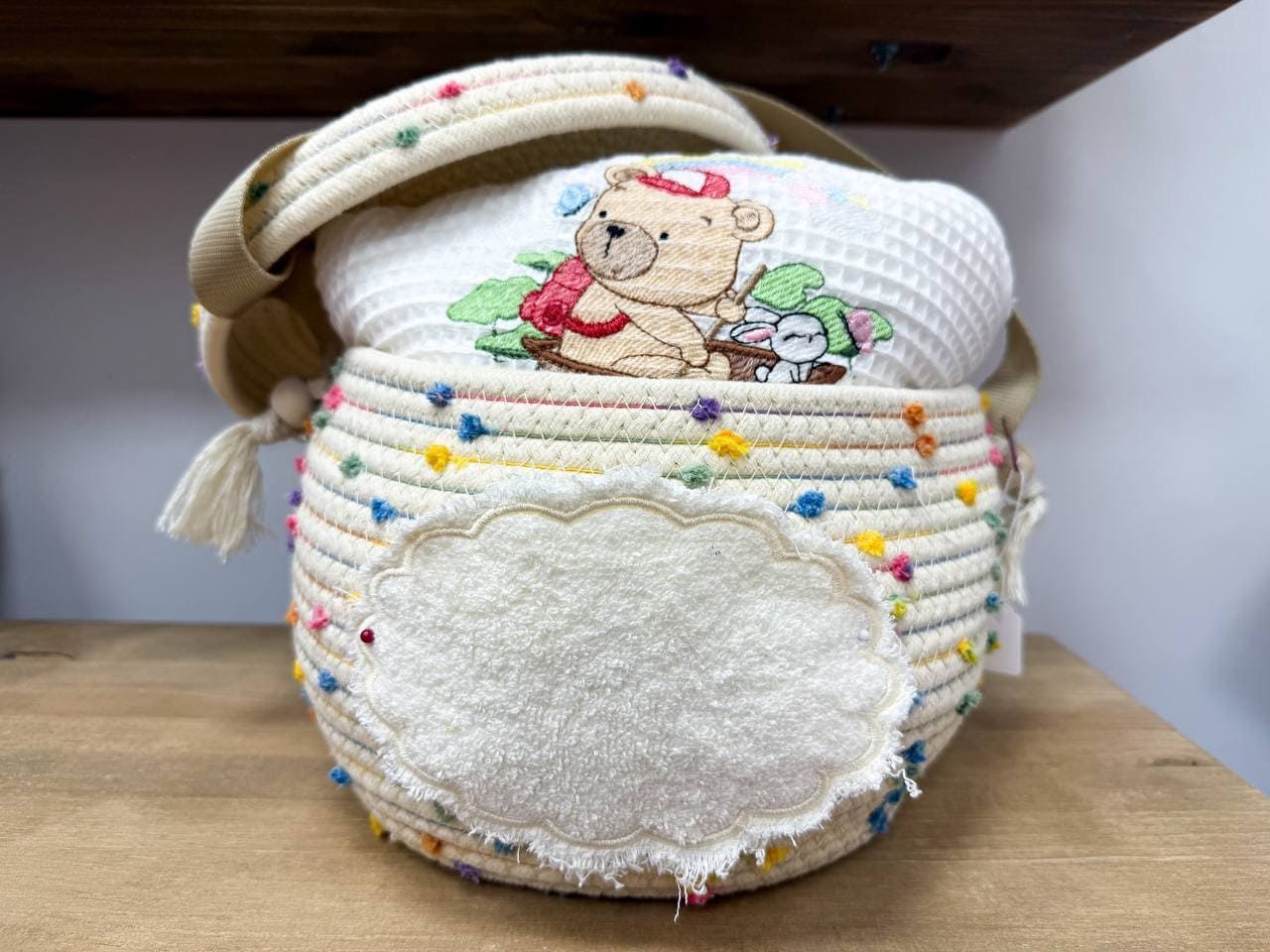 Una cesta con un osito de peluche bordado está sobre una mesa de madera.