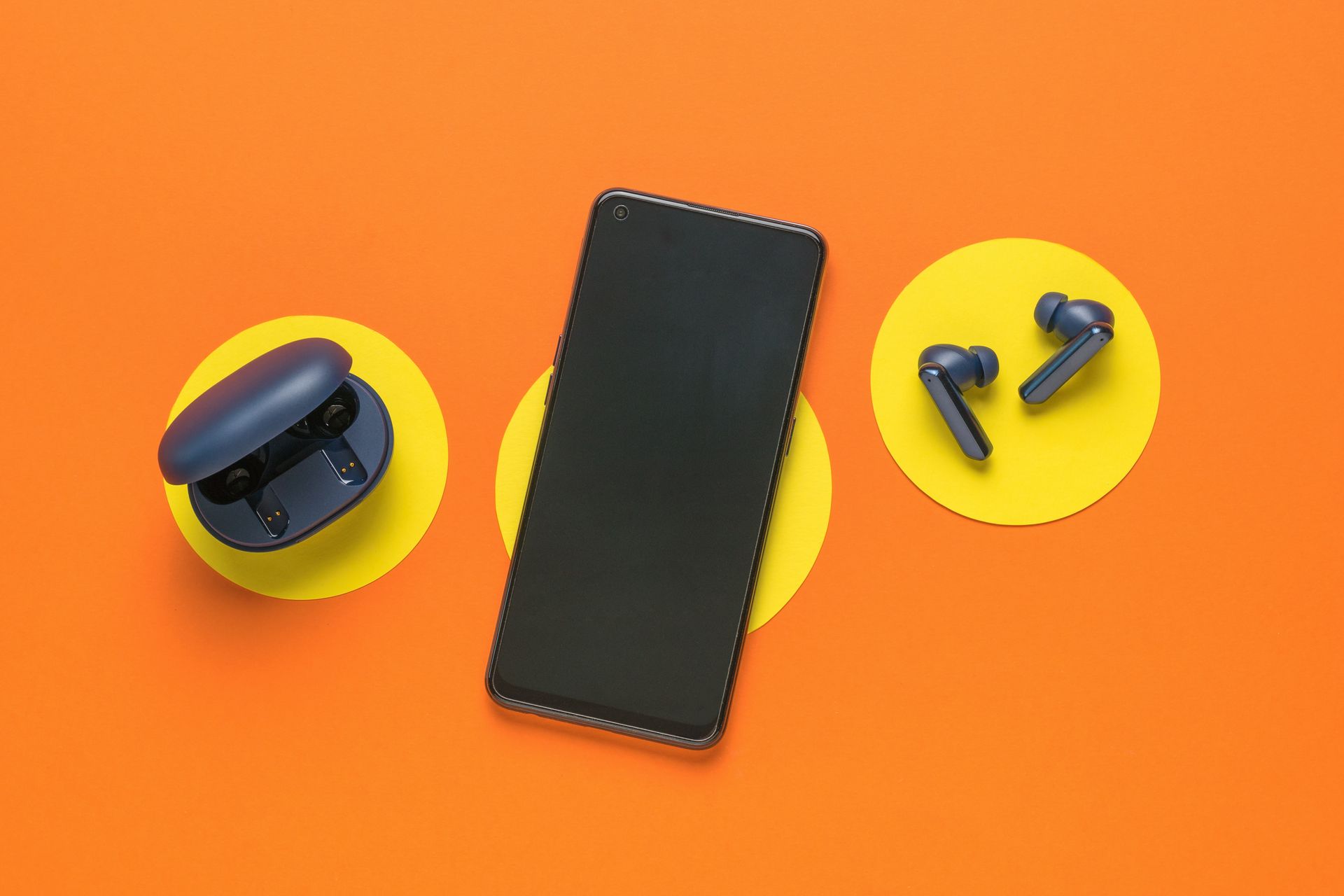 Smartphone noir avec écouteurs et étui de chargement sur des cercles jaunes sur fond orange.