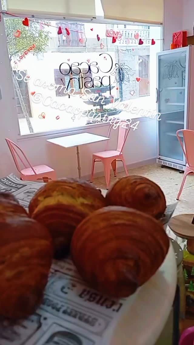 Cafetería-panadería con pasteles en primer plano, sillas rosas, mesas pequeñas y un escaparate luminoso.