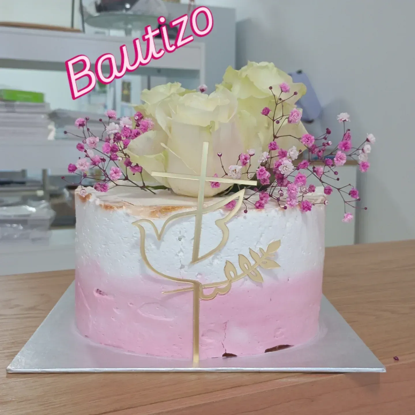 Tarta de bautizo rosa y blanca con flores, cruz dorada y adorno de “Bautizo” sobre una base plateada.