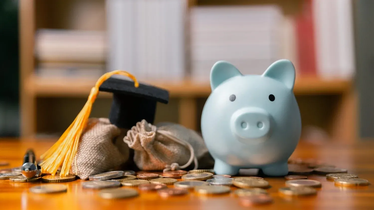 Una hucha azul claro se encuentra junto a un birrete de graduación, encima de dos pequeñas bolsas de tela rodeadas de monedas esparcidas.