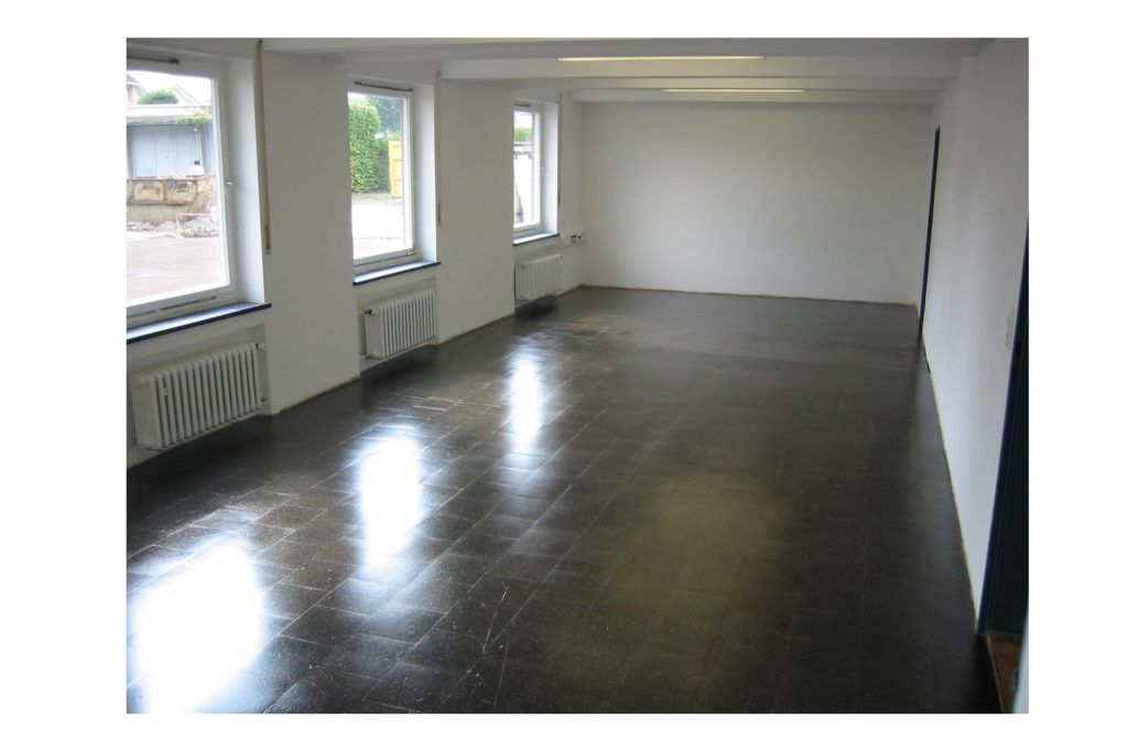 Leerer Raum mit glänzend dunklem Boden, weißen Wänden und Fenstern an der linken Seite.