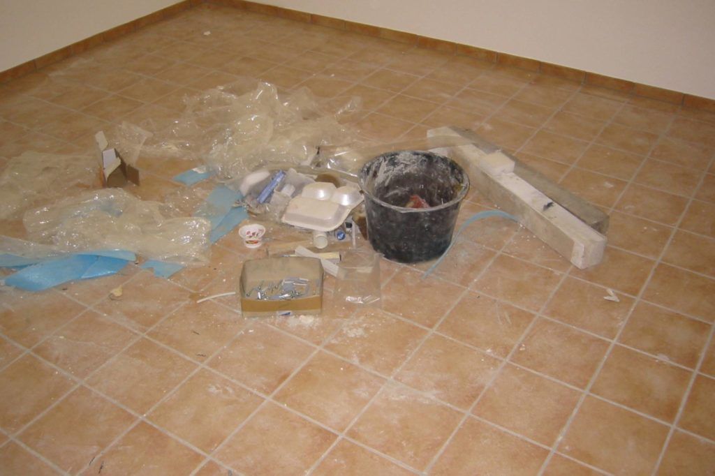 Bauschutt auf einem gefliesten Boden neben einem schwarzen Eimer und verstreuten Werkzeugen/Verpackungsmaterialien.