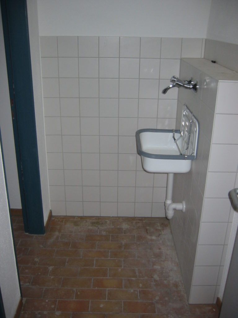 Kleine Toilette mit wandmontiertem Waschbecken, weiß gefliesten Wänden und Ziegelboden.