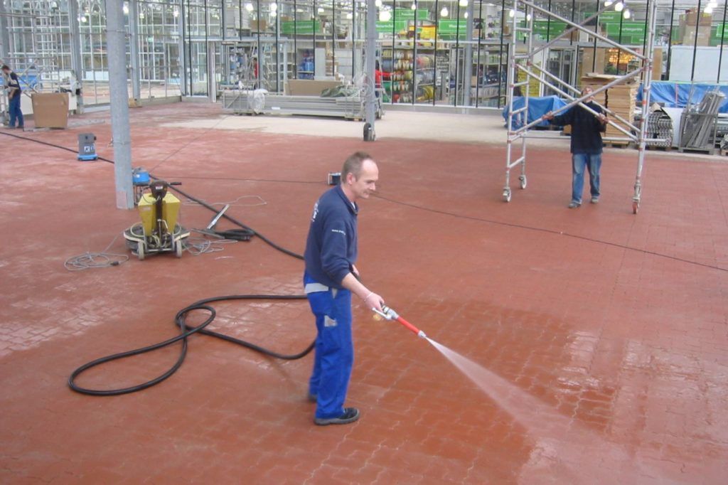 Ein Arbeiter in blauem Overall reinigt mit einem Hochdruckreiniger einen roten Backsteinboden in einem Industriegebiet.
