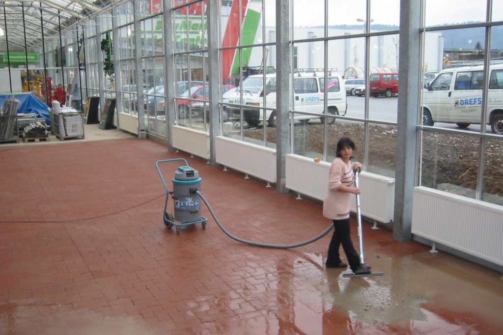 Ein Kind bespritzt drinnen mit einem Gartenschlauch einen nassen Boden und draußen parkende Autos.