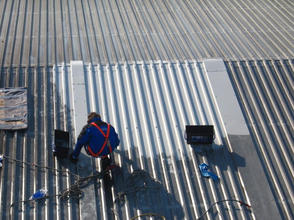 Ein Arbeiter arbeitet auf einem Wellblechdach und benutzt Werkzeuge in der Nähe eines schwarzen Eimers und Kabeln.