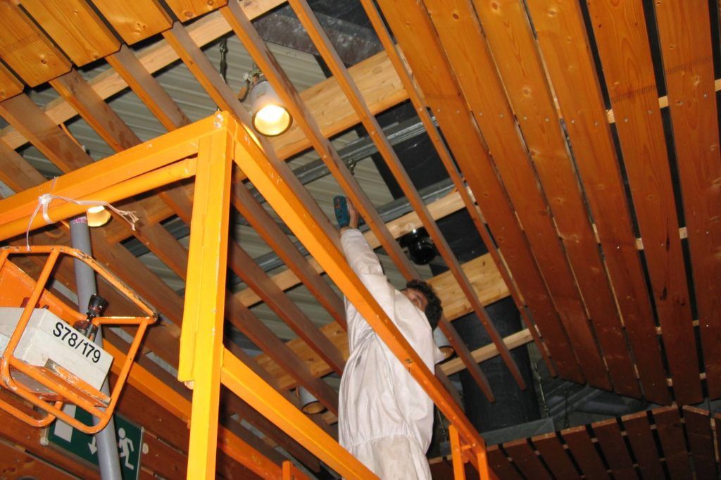Arbeiter montiert auf einem orangefarbenen Hubsteiger Holzdeckenplatten in einem im Bau befindlichen Raum.
