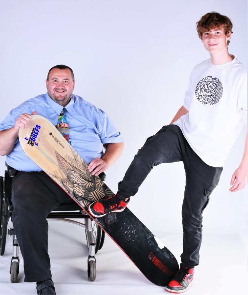 Zwei Personen posieren mit Skateboards; eine sitzt links in einem Rollstuhl, die andere steht rechts.