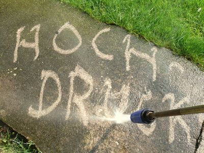 Kreideschrift auf dem Bürgersteig wird mit Wasser besprüht, Gras wächst am Rand.
