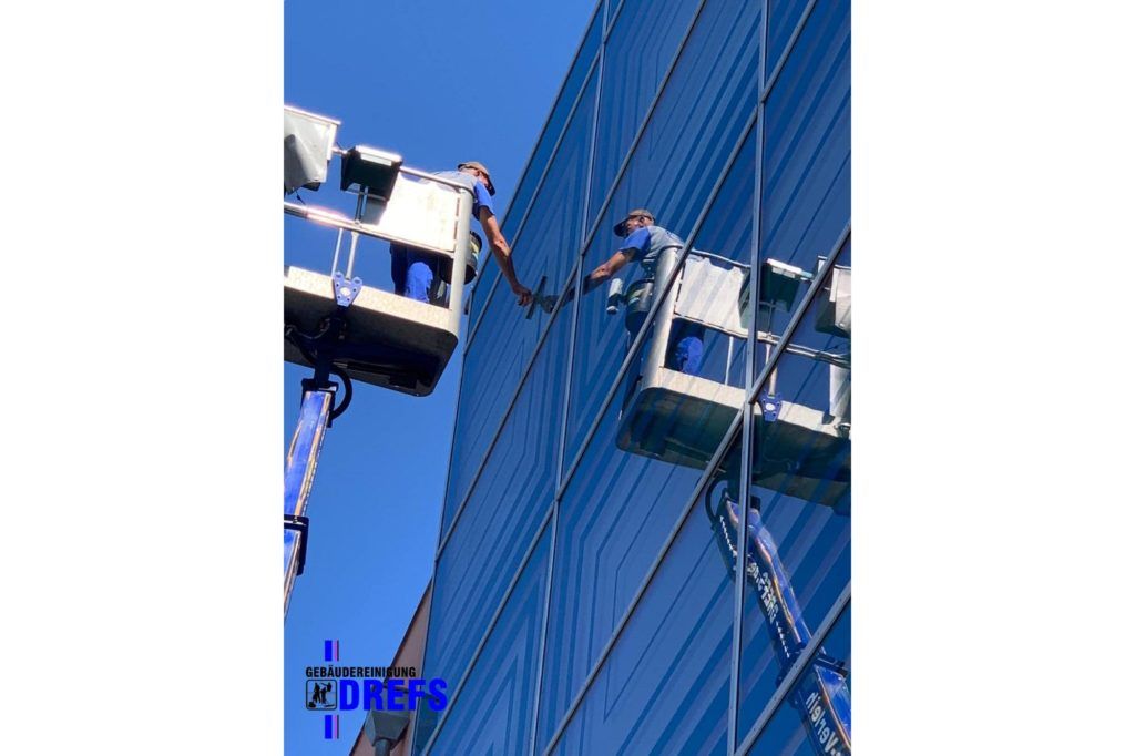 Zwei Arbeiter in Arbeitskörben reinigen die Fassade eines blauen Glaswolkenkratzers.