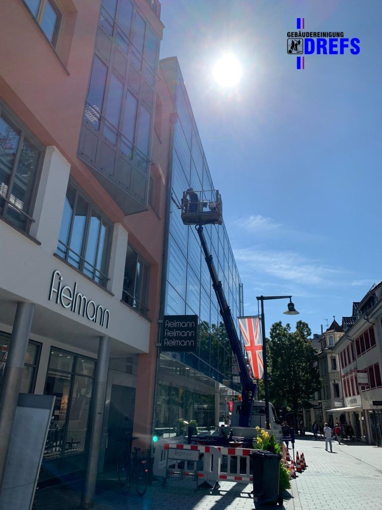 Sonnige Stadtstraße mit modernen Ladenfronten, einer Caféterrasse und Fußgängern entlang eines Gebäudes mit Glasfassade.