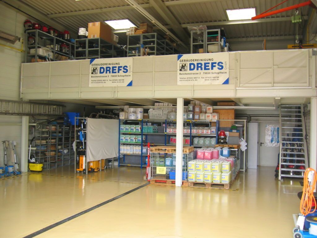 Industriehalle mit Regalen voller Vorräte, gestapelten Kisten und DREFS-Schildern auf einer Zwischenebene.