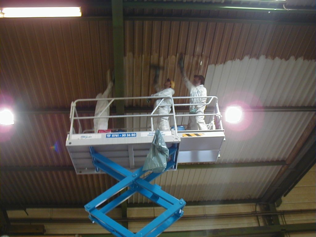 Zwei Arbeiter inspizieren auf einer blauen Scherenbühne die Decke einer Lagerhalle unter hellem Scheinwerferlicht.