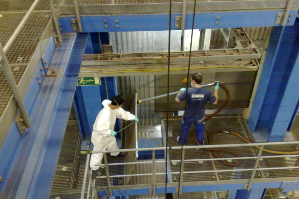 Zwei Arbeiter in Schutzkleidung reinigen einen großen Industrietank oder eine Grube mit Schläuchen und Werkzeugen.