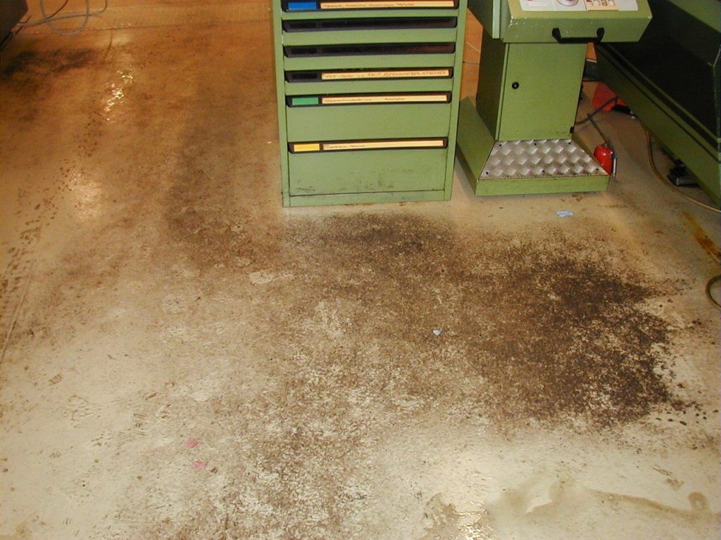 Schmutziger Betonboden in der Werkstatt neben grünen Werkzeugschränken und Maschinen, mit Ölflecken und verstreutem Schutt
