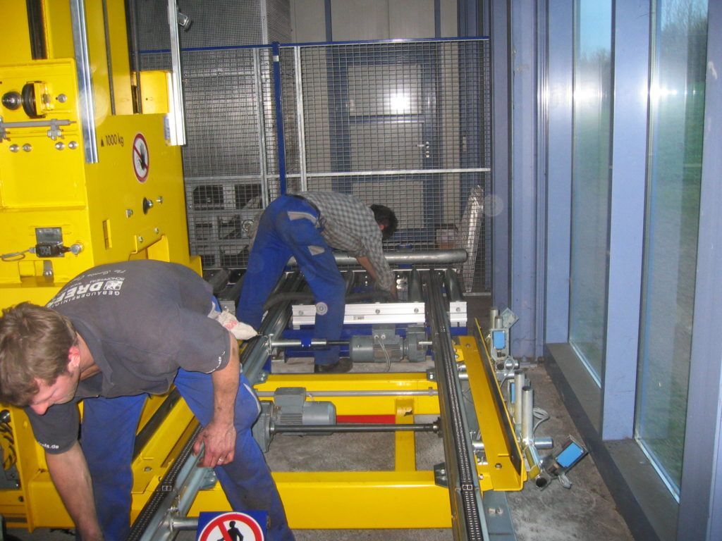 Arbeiter bedienen gelbe Maschinen in einer Industrieanlage hinter einem Sicherheitskäfig.