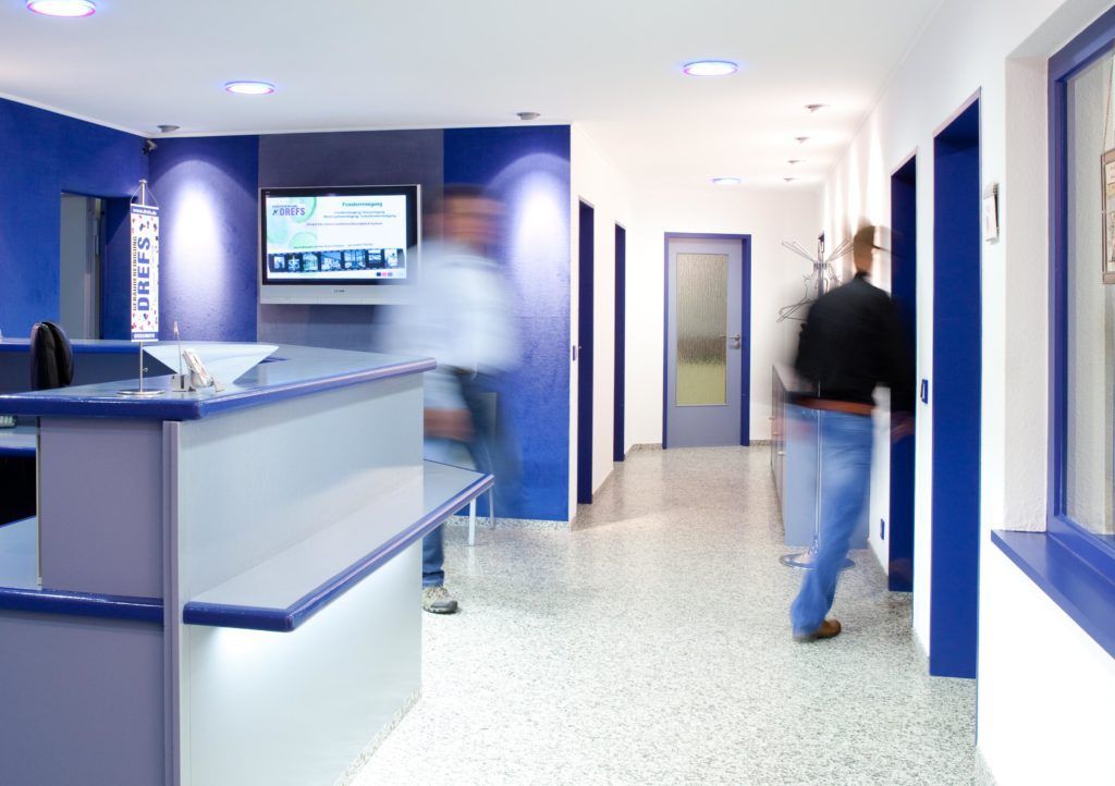 Ein heller Krankenhausflur, in dem zwei verschwommene Personen an blauen Wänden und einem Empfangstresen vorbeigehen.