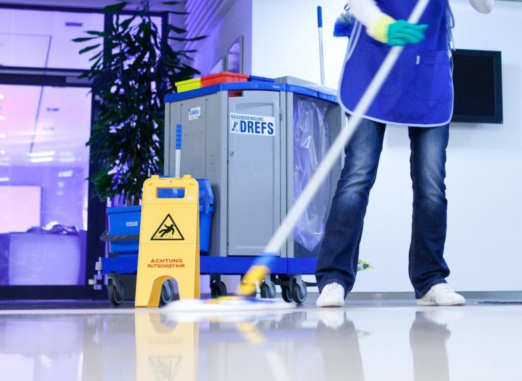Ein Hausmeister wischt einen glänzenden Büroboden neben einem gelben Warnschild für nassen Boden und einem Reinigungswagen.