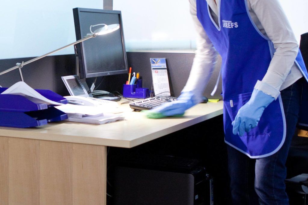 Eine Person, die Handschuhe trägt, reinigt einen Büroschreibtisch mit einer Sprühflasche und einem Tuch.